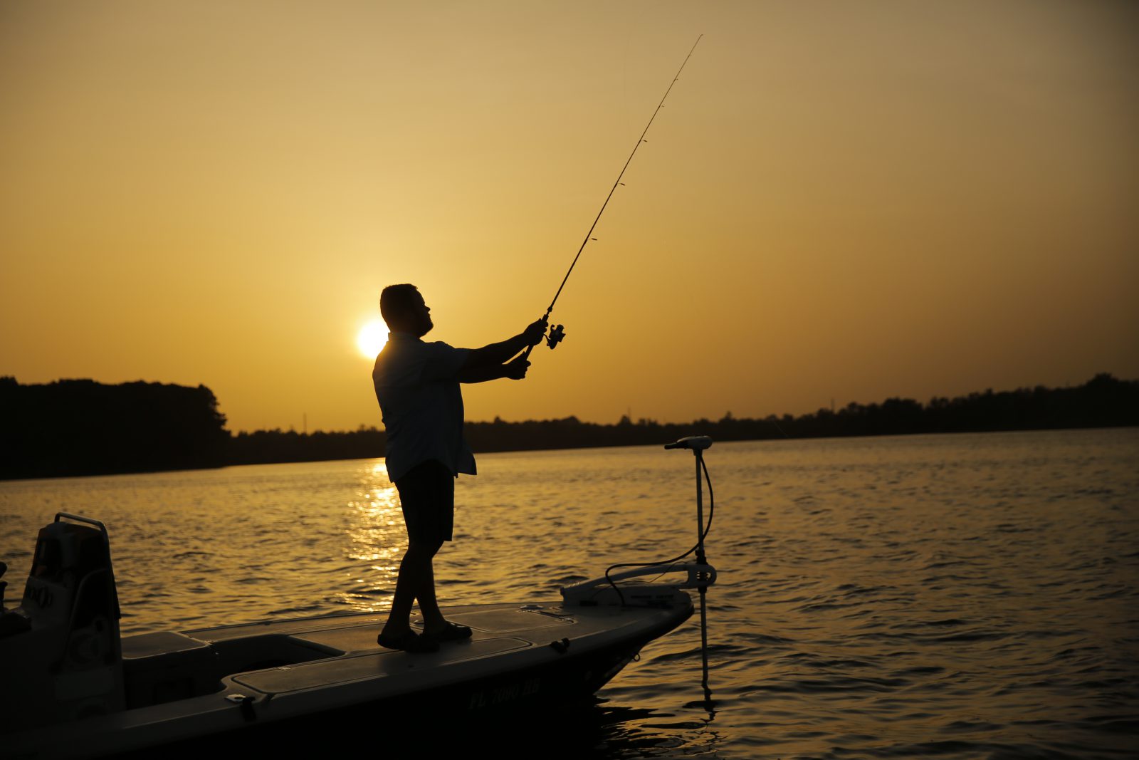 Man standing on deck of bass boat fishing in the middle of a lake as the sun is setting