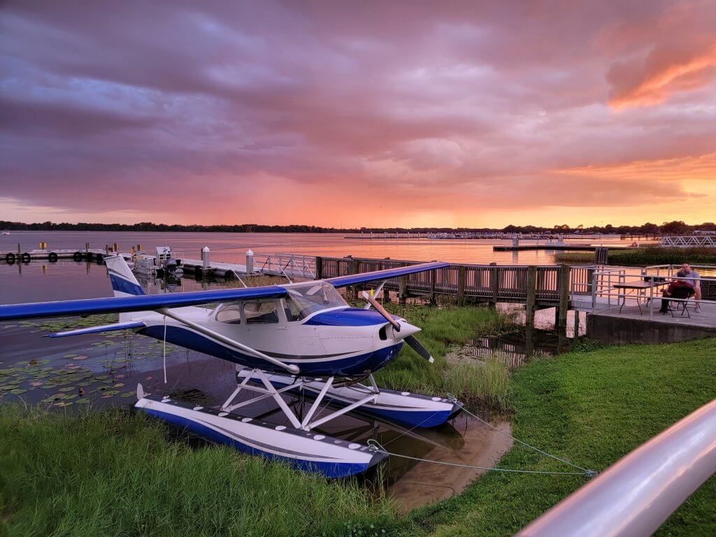 blue and white seaplane on grassy lake shore, purple and orange cloudy sunset in background