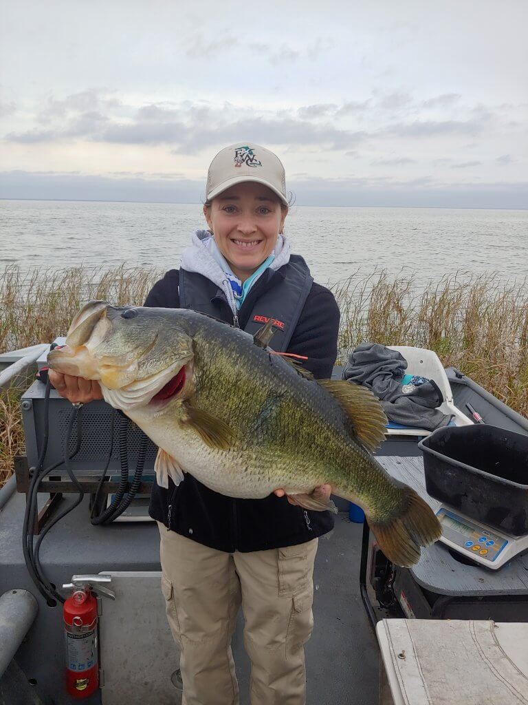Young lady holding a largemouth bass horizontally with a lake in the background