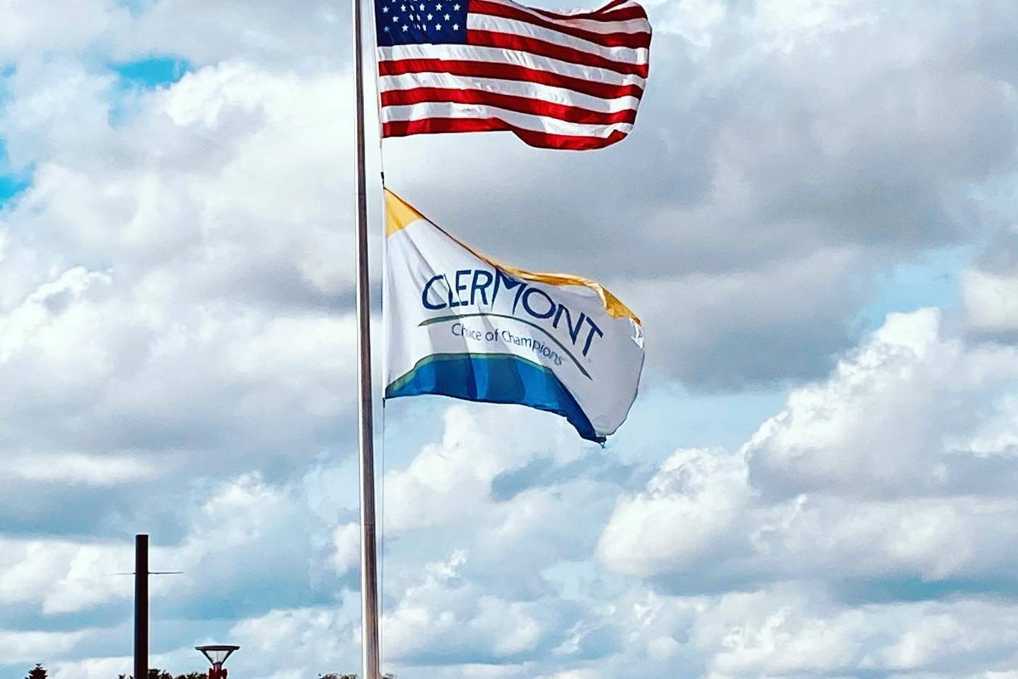 Photo of American flag above Clermont city flag against cloudy blue sky.