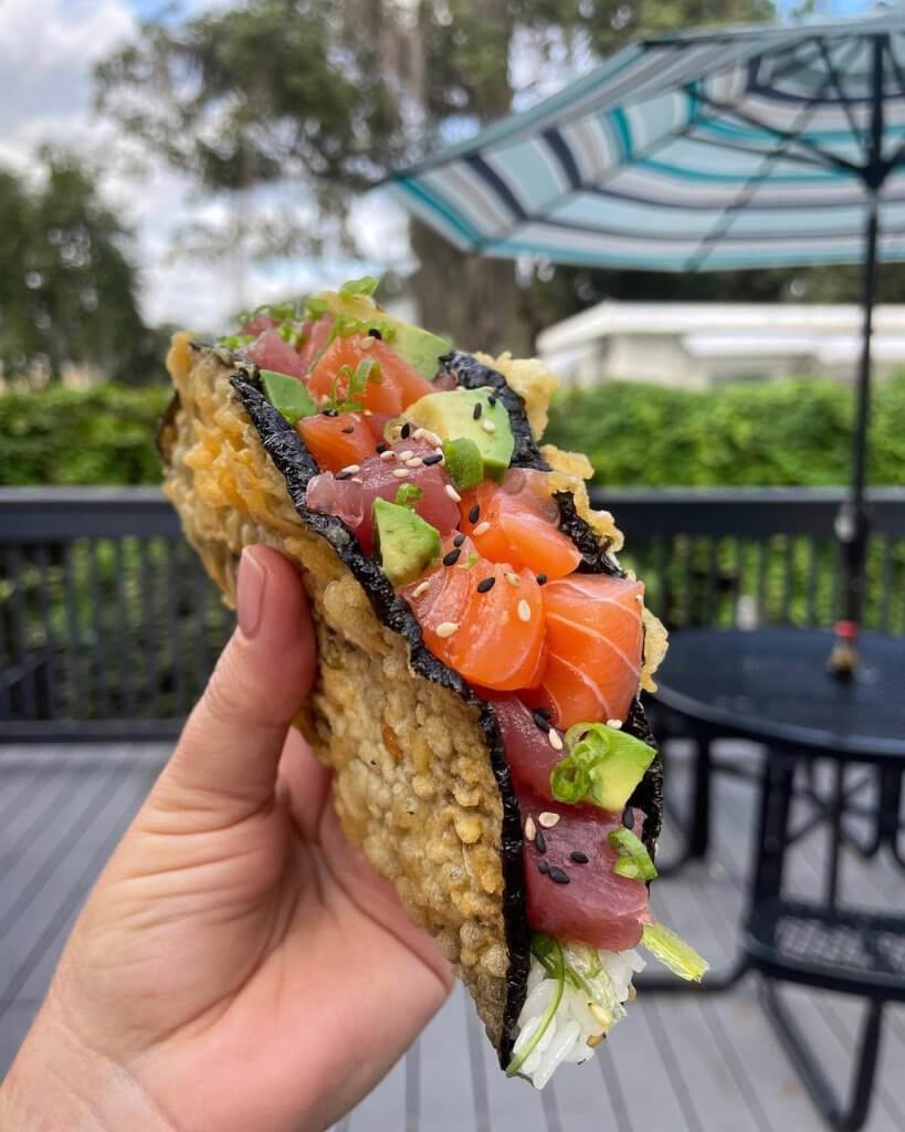 Hand holding sushi taco in foreground, outdoor patio in background